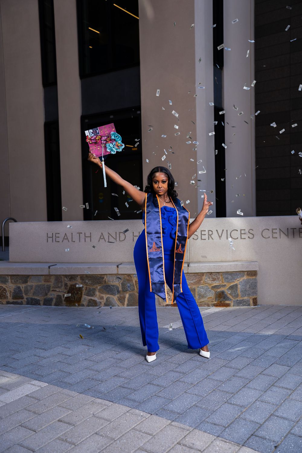 Graduate in blue cap and gown celebrates with confetti during outdoor graduation photo session at health services center.