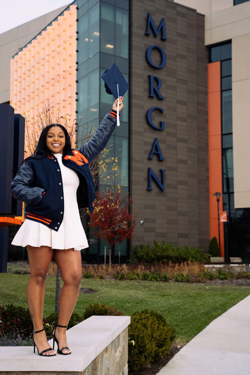 Joyful graduate in black and white attire celebrates achievement in front of university building.