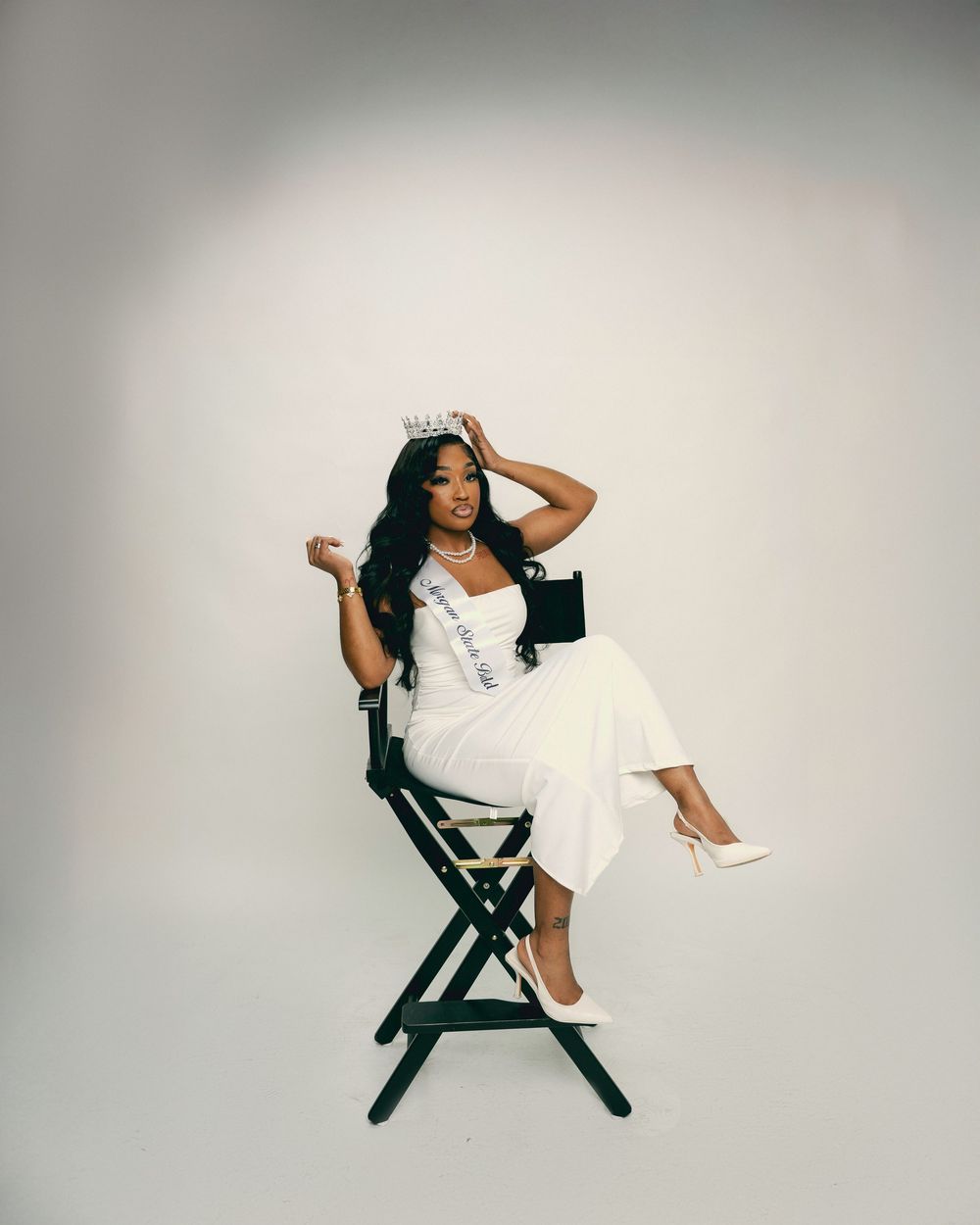 A person in a white dress poses dramatically across a director's chair against a light gray studio backdrop.