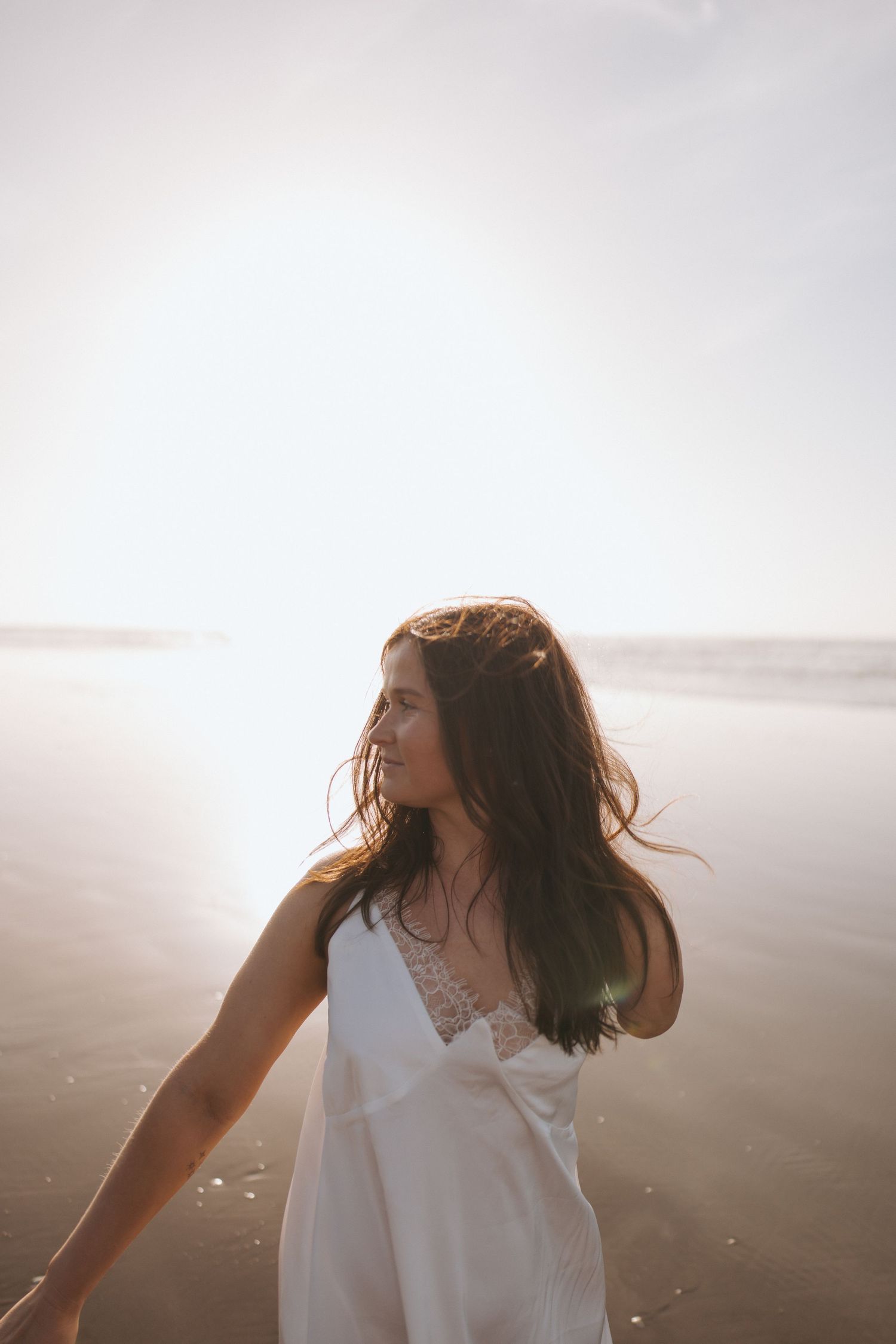 Young woman in white dress standing on beach at sunset in New Zealand.
