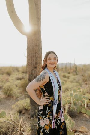 Graduate in floral dress poses near saguaro cactus at sunset.