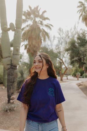 Person wearing navy blue t-shirt walks along desert path with saguaro cacti and palm trees.