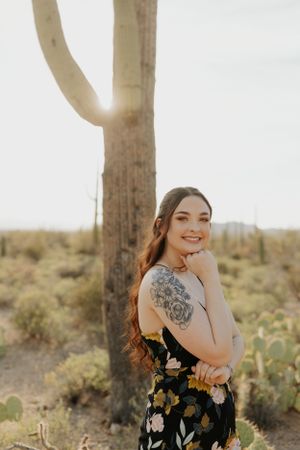 A figure in a black floral dress poses expressively among desert cacti during golden hour.