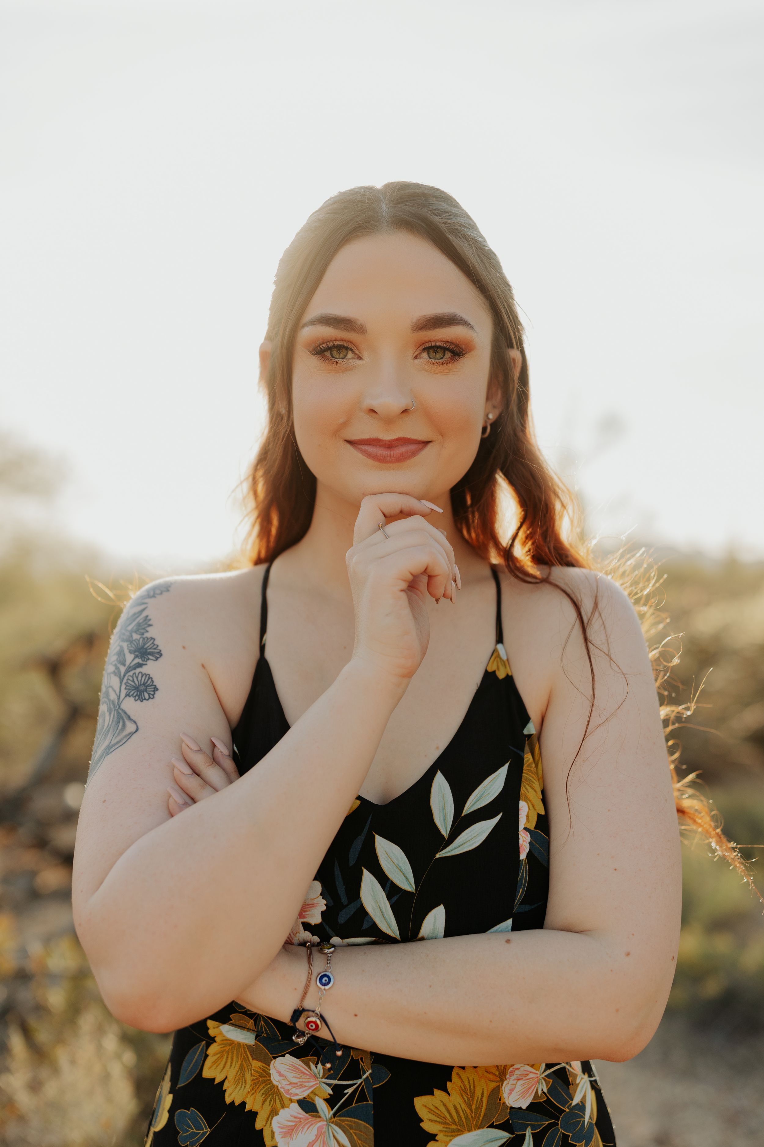 Graduate in floral dress poses with graduation stole against desert background.