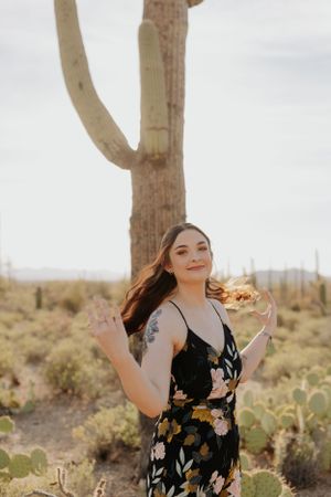 Graduate in floral dress poses playfully near saguaro cactus at sunset.