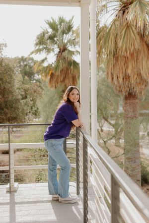 Person in purple shirt and jeans poses playfully against white railing with palm trees in background.