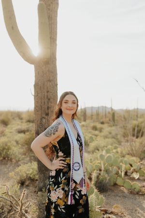 Graduate in floral dress and stole poses against desert landscape with saguaro cacti.