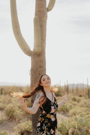 A floral dress-clad figure poses playfully against a desert landscape with saguaro cacti at sunset.
