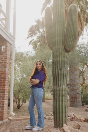 Person in blue shirt poses cheerfully next to large saguaro cactus and brick columns.