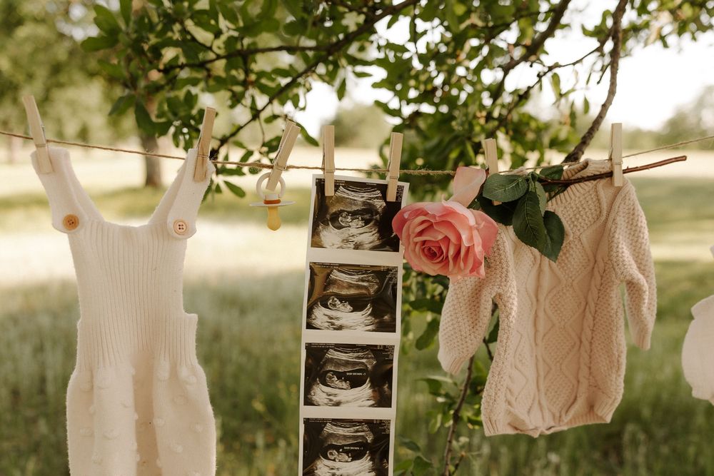 Baby clothes and ultrasound photos hang on a clothesline against a soft natural outdoor backdrop.