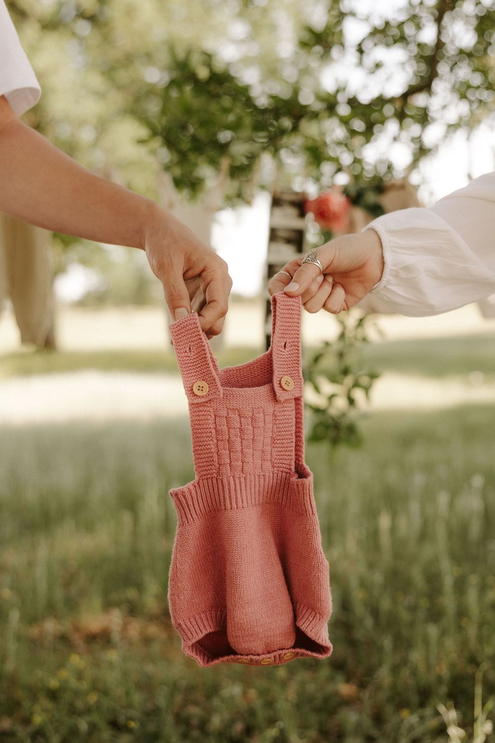 A small pink knitted baby romper being held up against a blurred natural outdoor background.