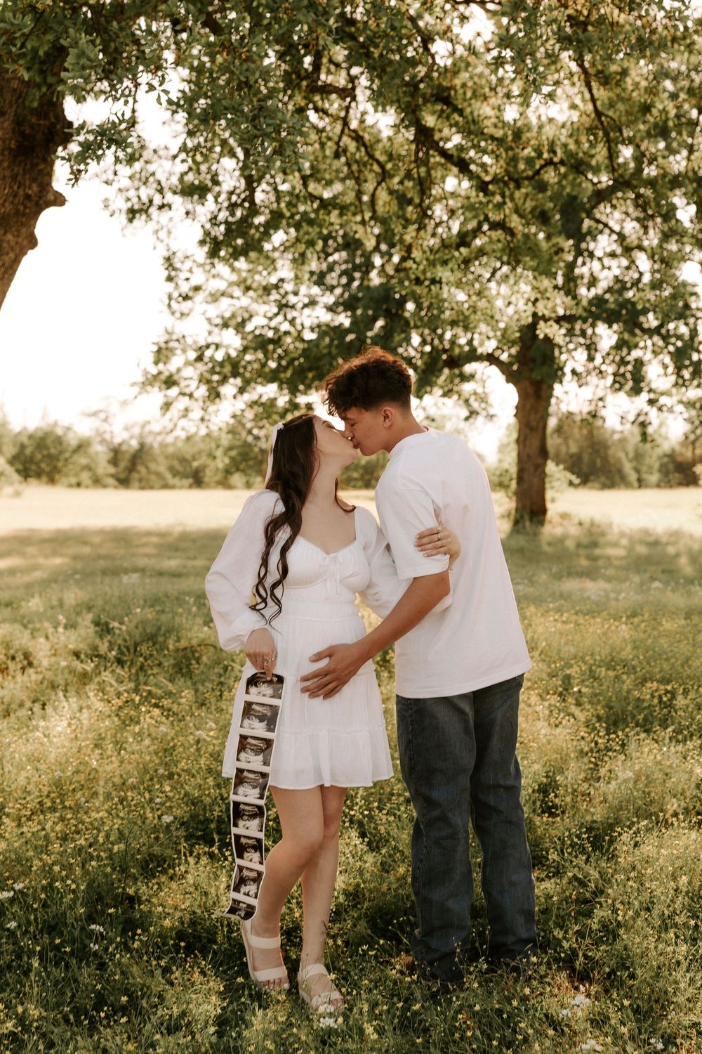 Couple holding ultrasound photos while standing in a sunlit meadow with golden wildflowers and trees.
