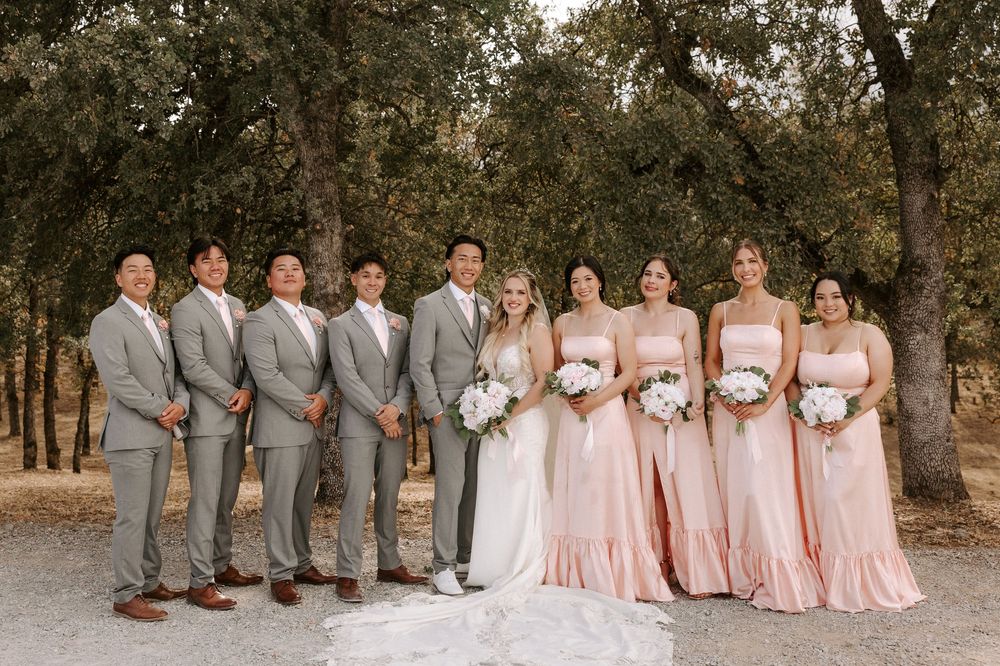 Wedding party in pink dresses and gray suits poses together for formal photos outdoors under trees.