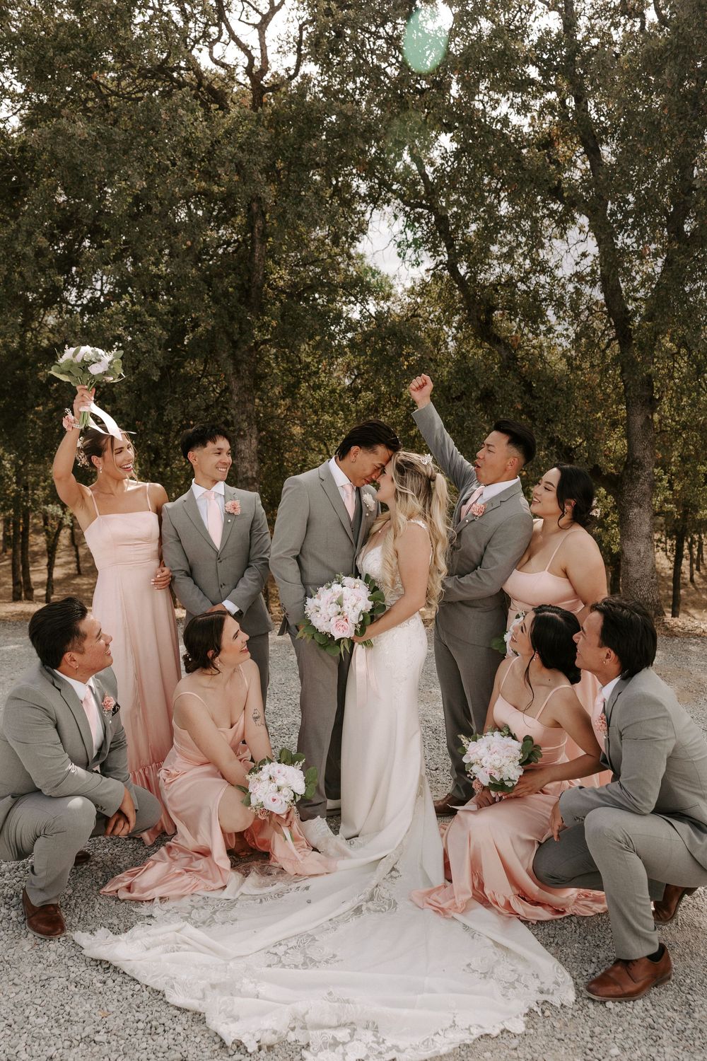 Wedding party in blush pink dresses and gray suits pose together outdoors under trees on a gravel path.
