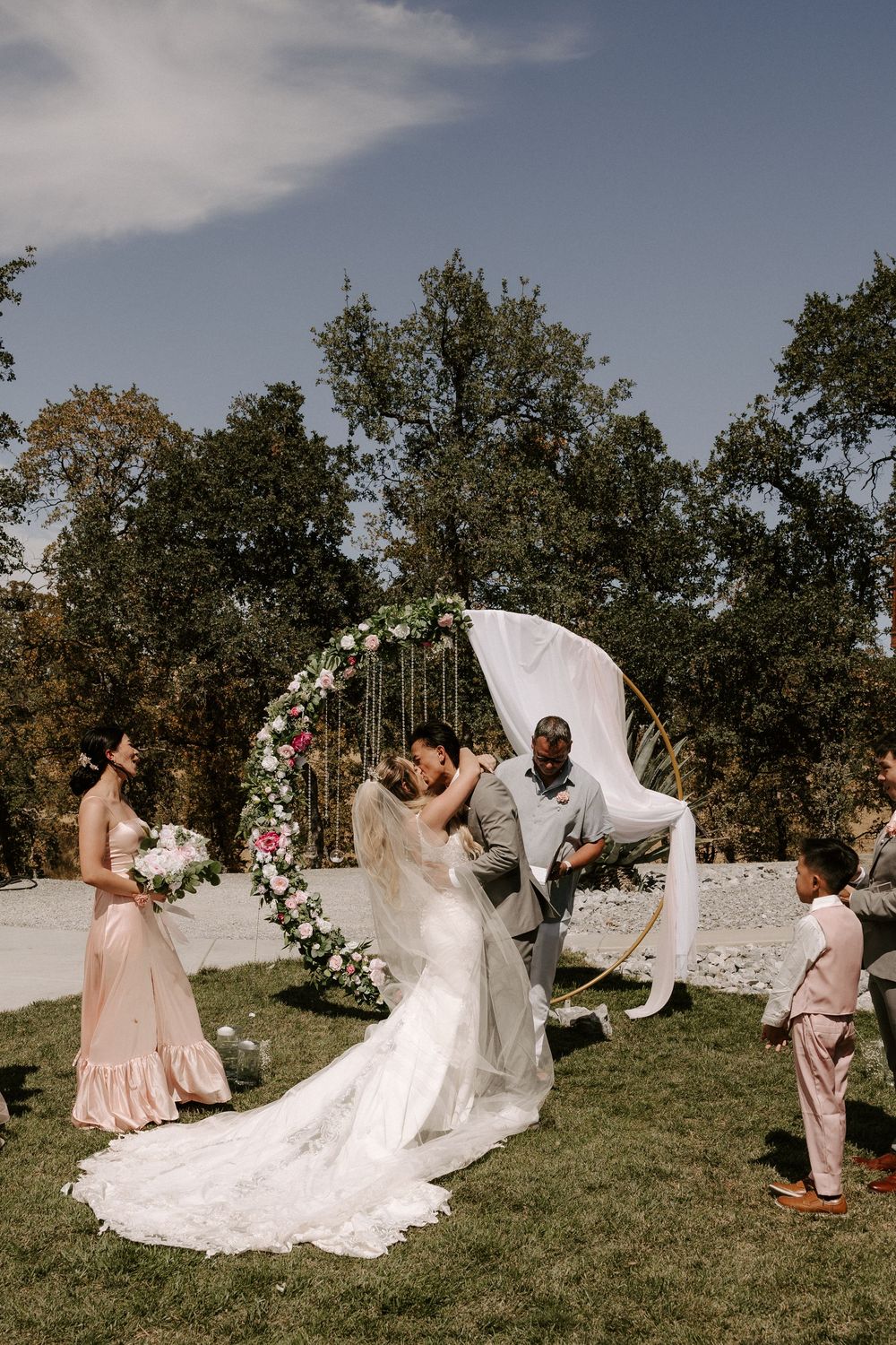 Wedding party having fun with flowing veil on a grassy hill with trees in the background.