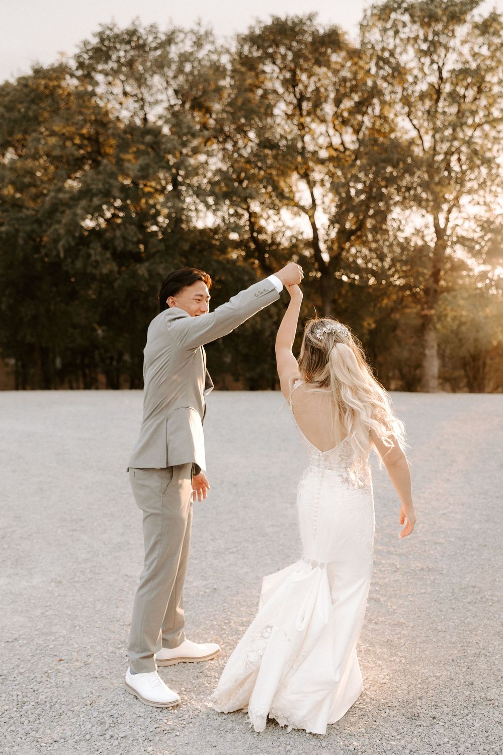 A couple in wedding attire dances joyfully together on a gravel path at sunset with trees in the background.