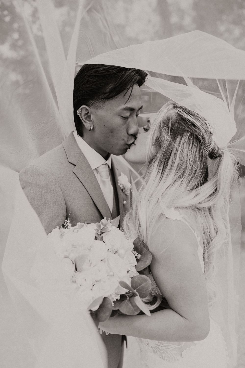 A romantic black and white wedding photo showing a tender kiss between newlyweds under a flowing veil.