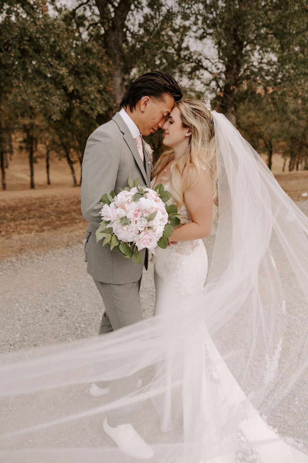Romantic wedding couple embraces outdoors in a rustic setting with flowing white veil and pink floral bouquet.
