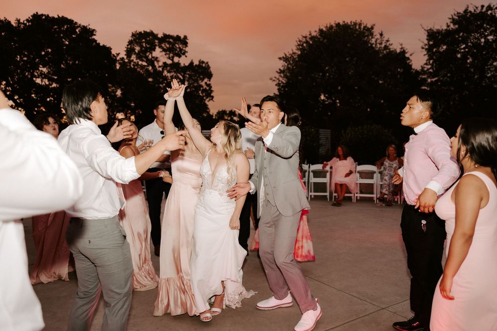 Wedding guests dance and celebrate together during an outdoor reception at sunset with pink sky.