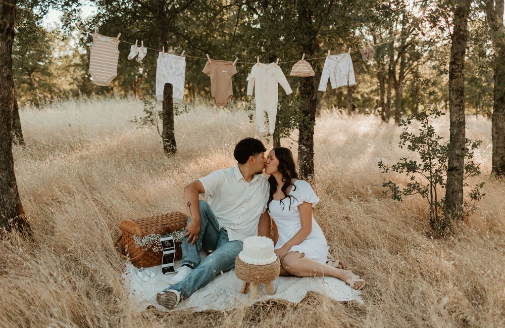 A couple shares a romantic moment while sitting in tall golden grass during sunset in a wooded area.