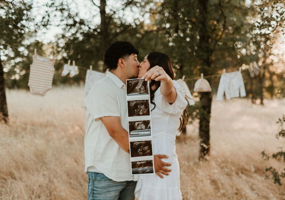 A couple embraces in a golden field at sunset while holding ultrasound photos.