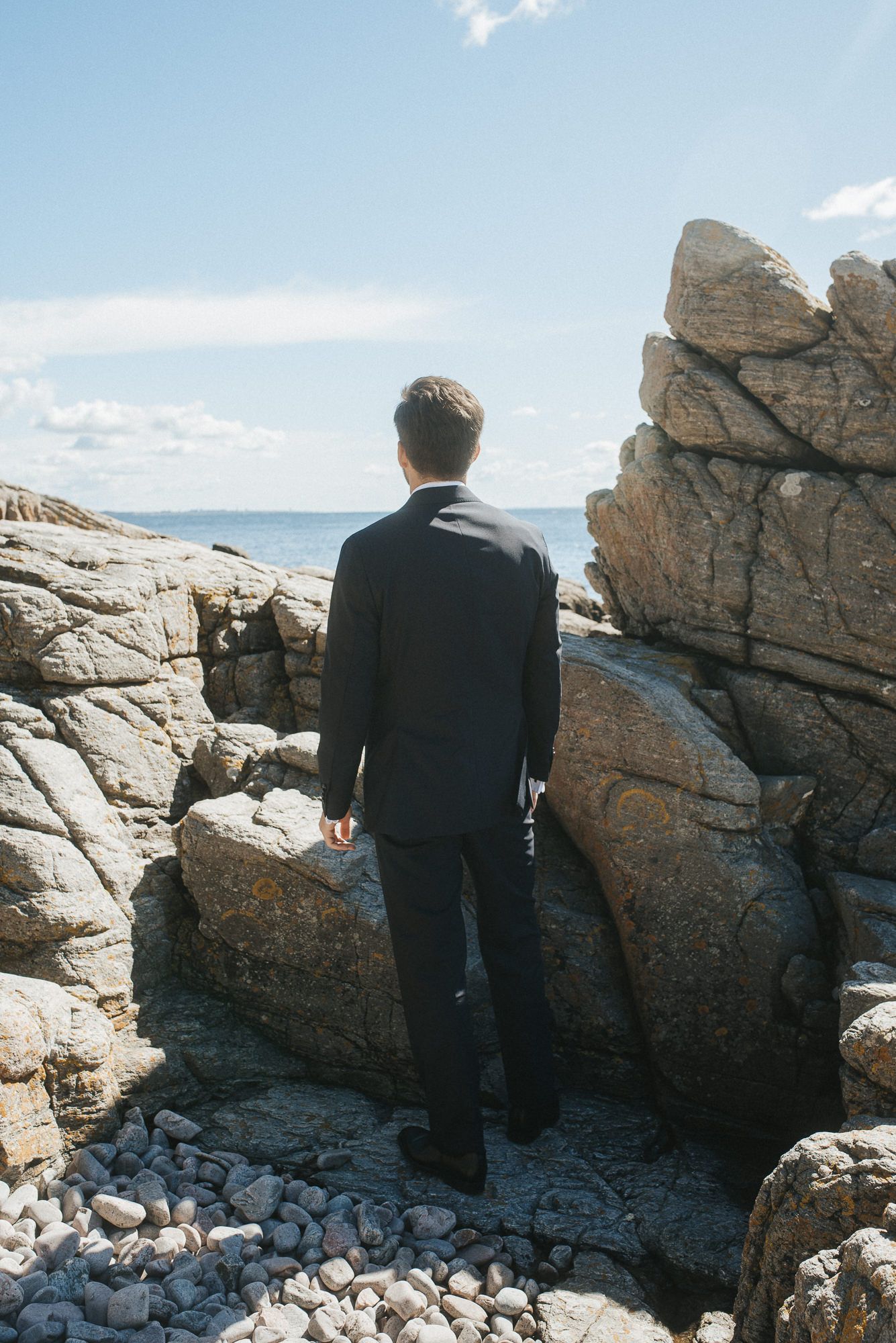 A person in dark clothing sits contemplatively on rocky coastal cliffs overlooking the ocean on a bright sunny day.