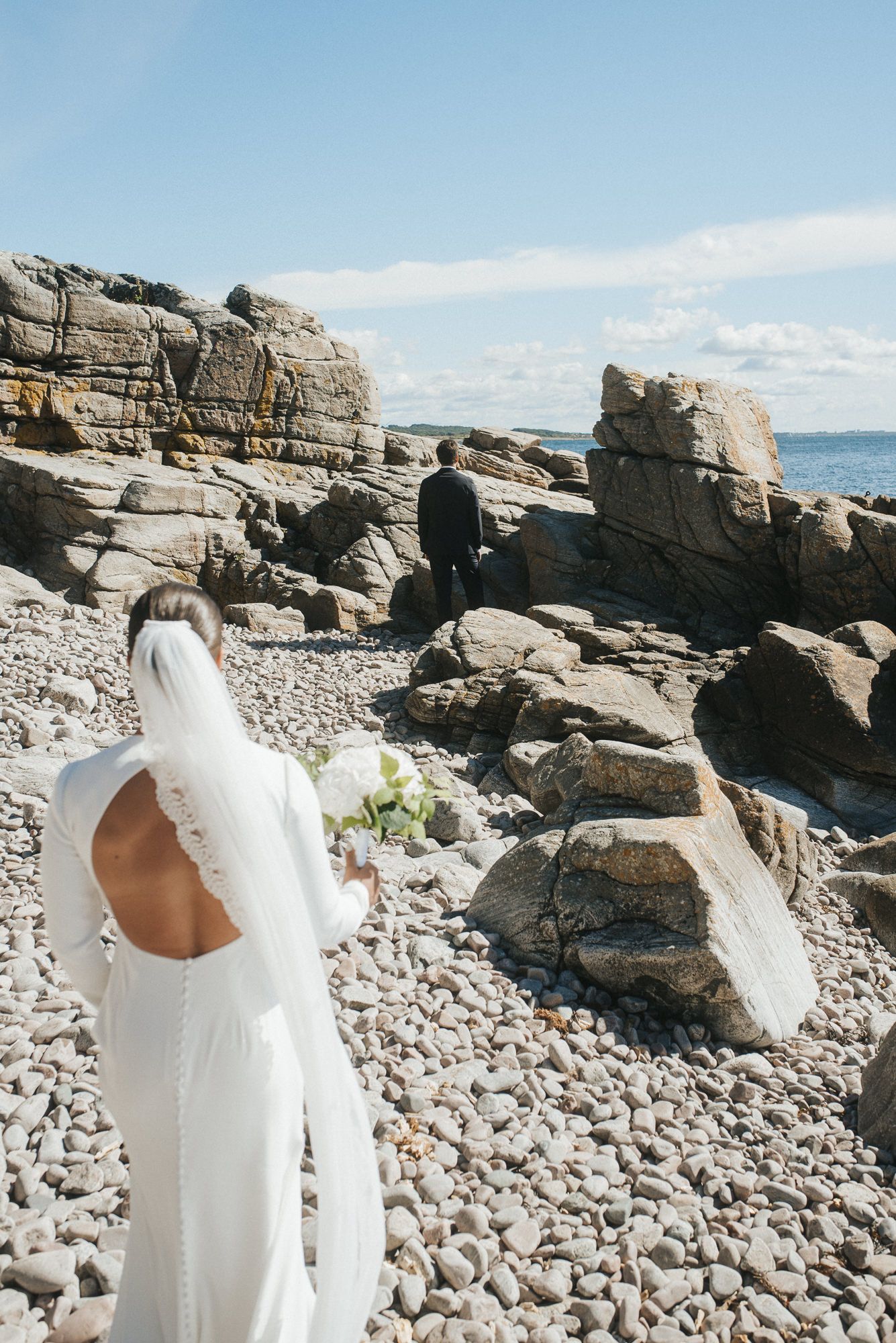 A wedding dress detail shot showing the back cutout design against a rocky coastal backdrop.