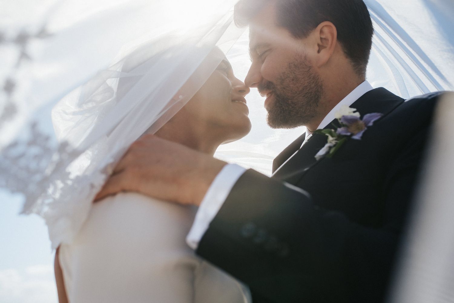 A romantic wedding couple embraces and kisses in front of a bright sunlit background creating a dreamy silhouette.