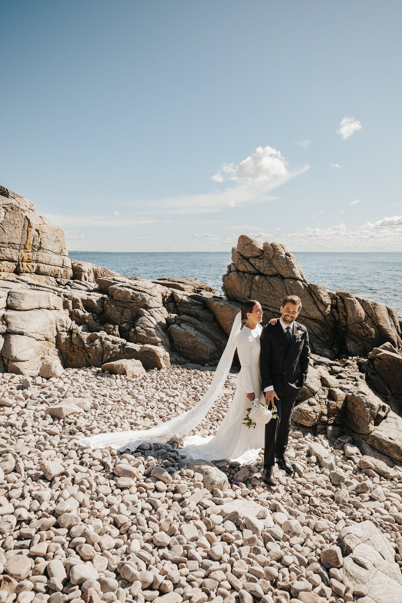 A couple poses on rocky coastal cliffs during their wedding photoshoot on a sunny day by the ocean.