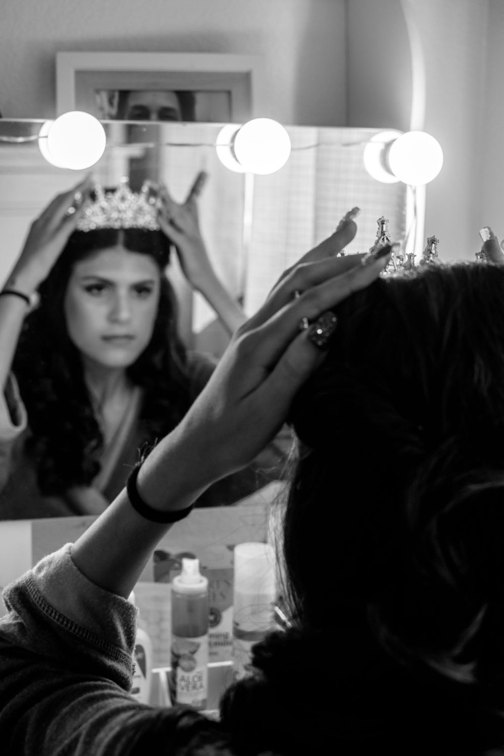 Black and white image of someone adjusting a delicate crown or tiara in a mirror with vanity lighting.