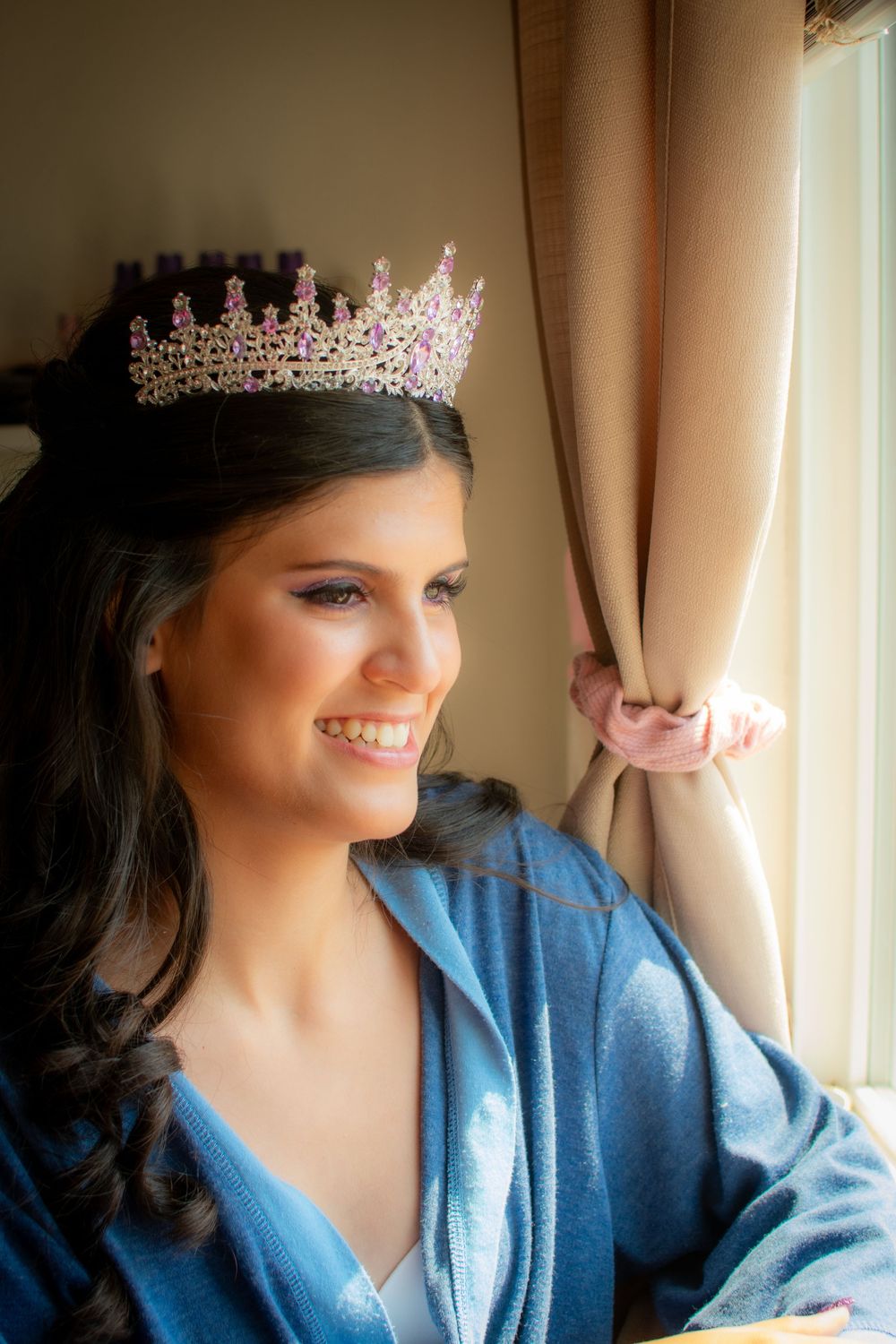 Person in blue robe wearing sparkly crown poses near window with soft lighting and curtains.