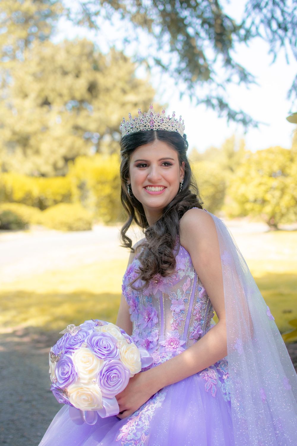 Quinceañera celebration in a park featuring a lavender gown, tiara and floral bouquet in soft natural lighting.