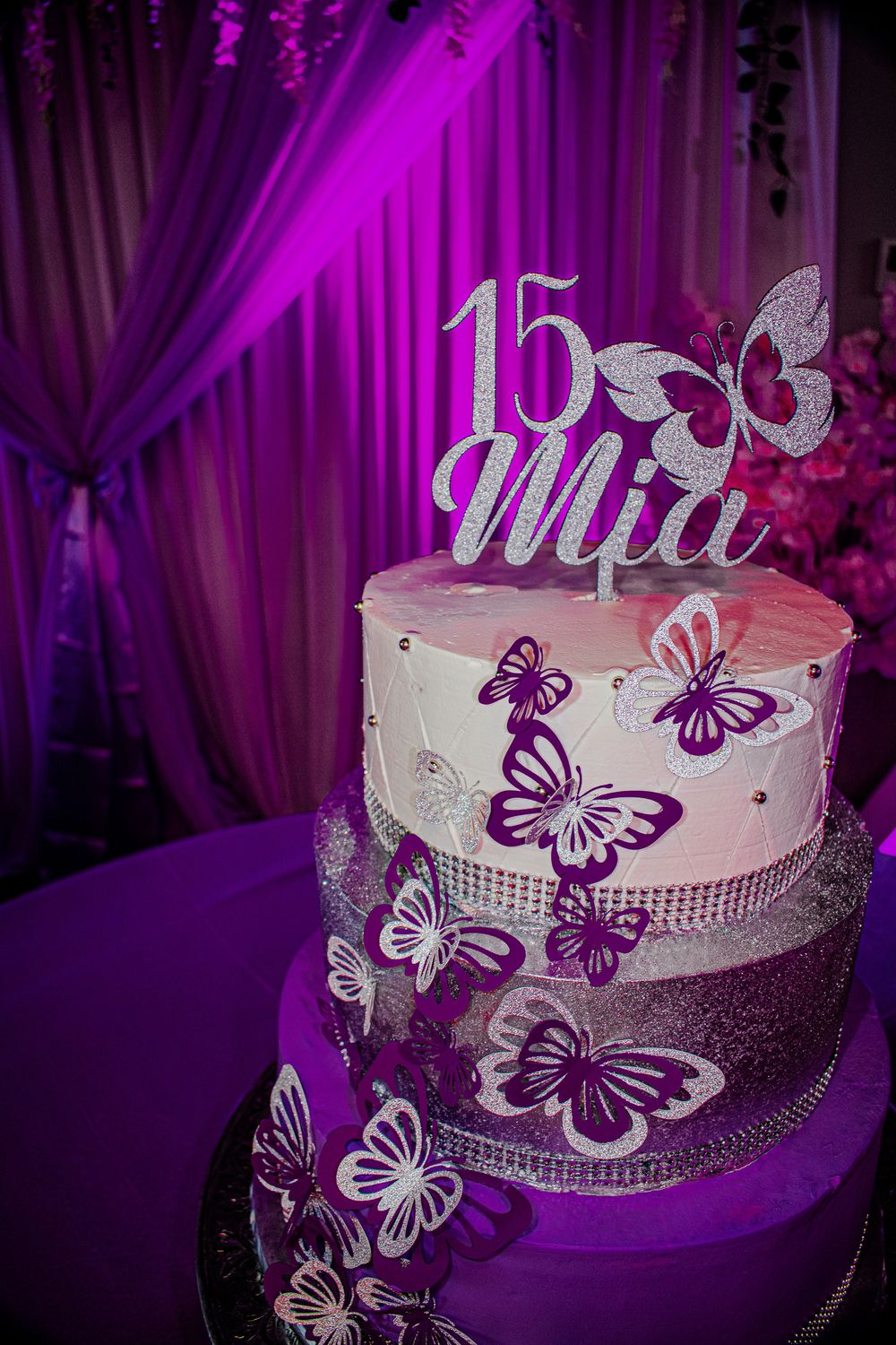 A three-tiered Quinceanera cake decorated with purple butterflies and a sparkly '15' topper under purple lighting.