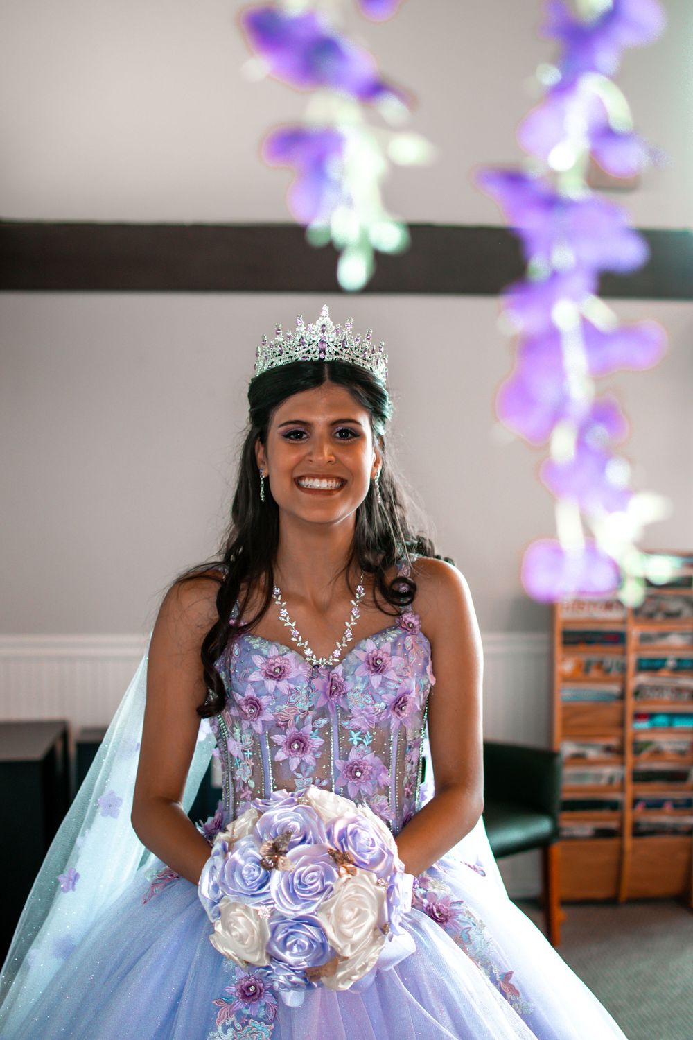 A celebrant in a lavender quinceañera gown holds a purple and white bouquet under floral decorations.