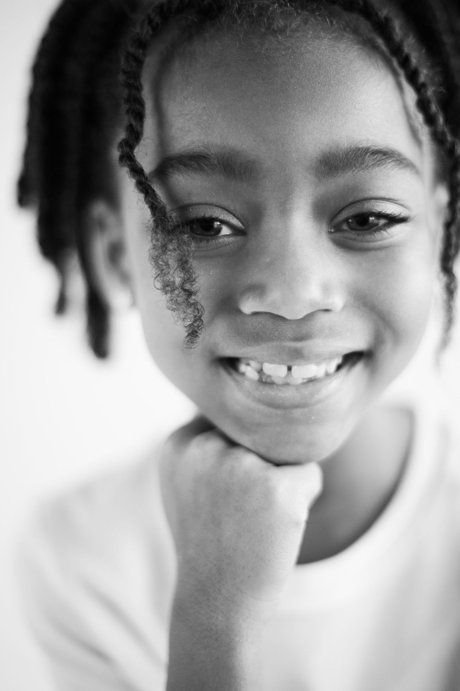 Black and white portrait of a child with braided hair smiling warmly while resting their chin on their hand.