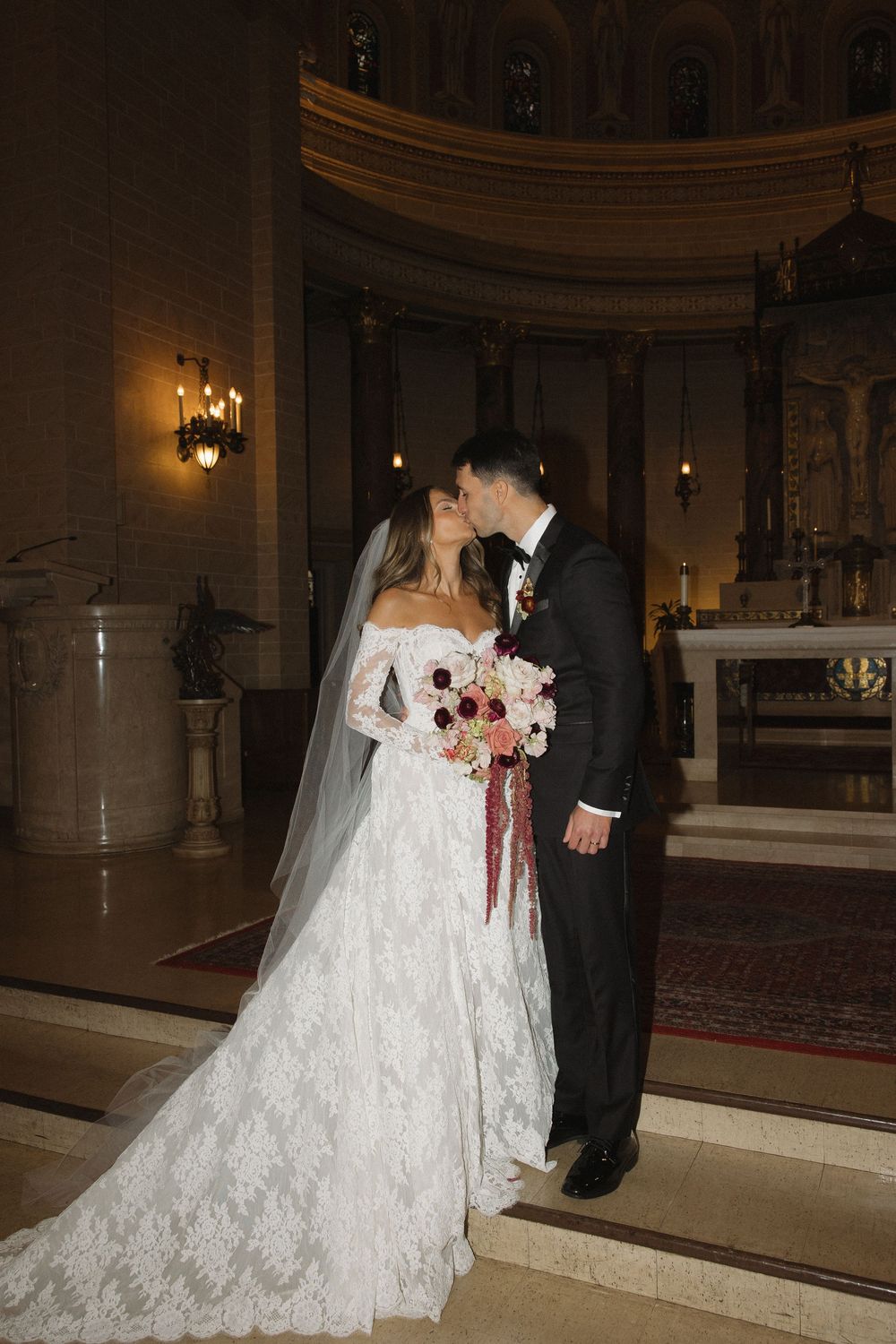 A bride in a lace wedding gown and groom in black tuxedo stand together in an ornate church sanctuary.