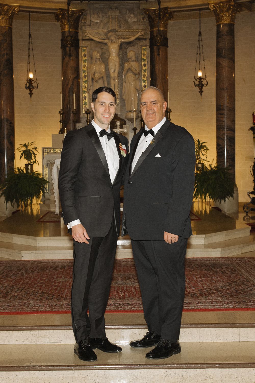 Two men in formal black tuxedos standing together in an elegant ballroom with ornate decor and wall sconces.