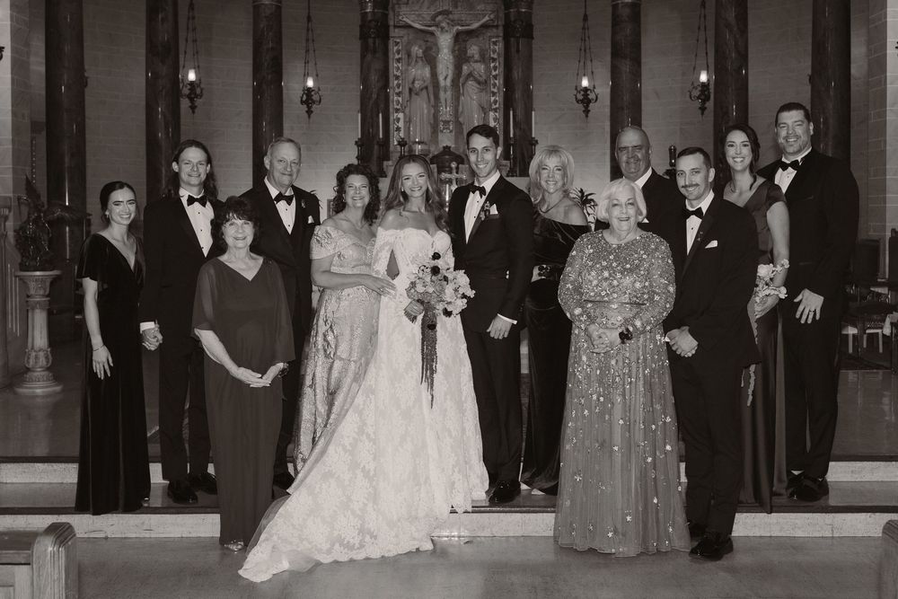 A black and white photograph shows a formal wedding party posing together in an elegant church or venue interior.