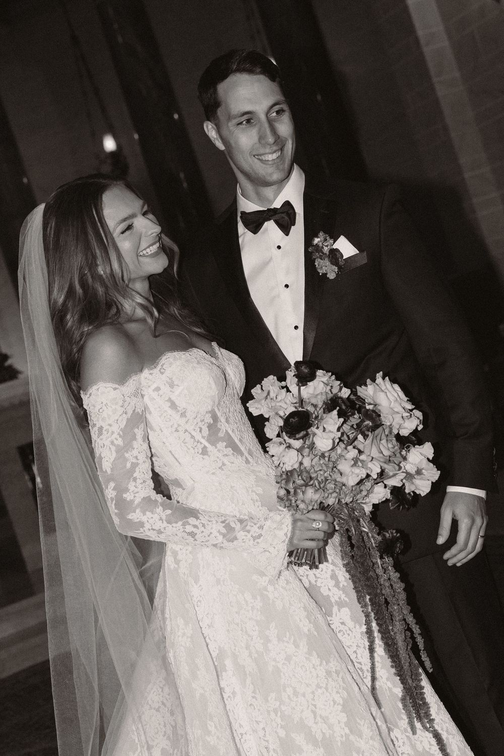 A happy newlywed couple shares a candid moment together while the bride holds a beautiful floral bouquet.