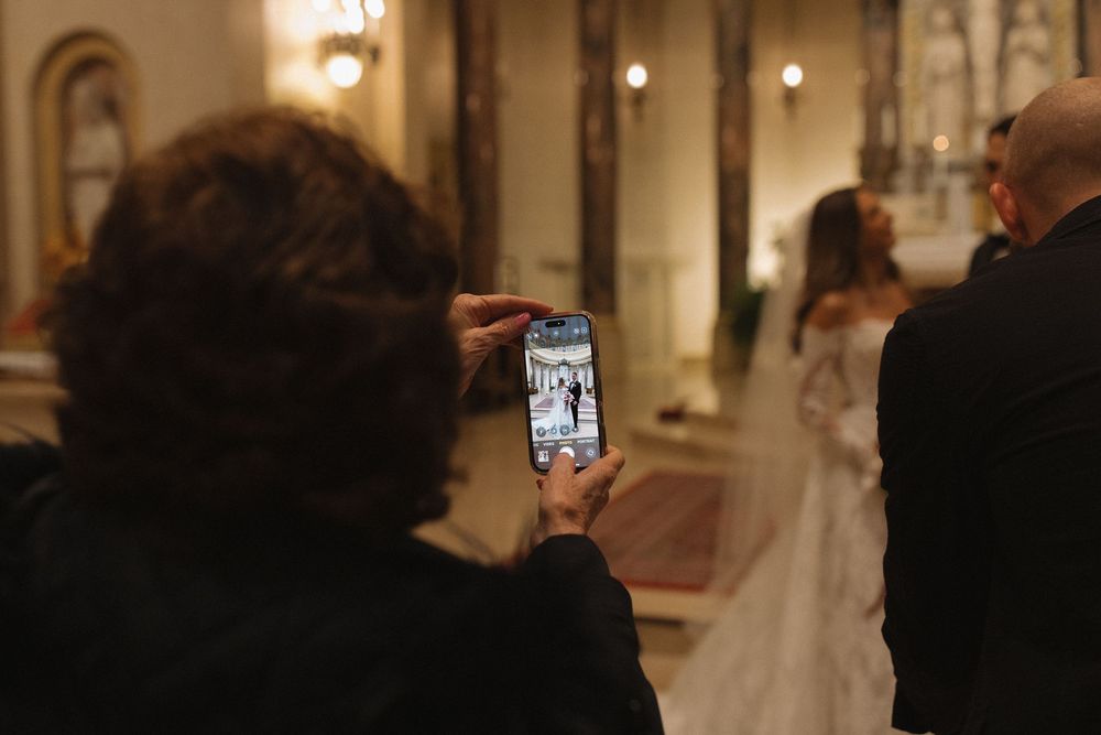 A wedding guest takes a photo on their phone of the bride standing in an ornate church aisle.