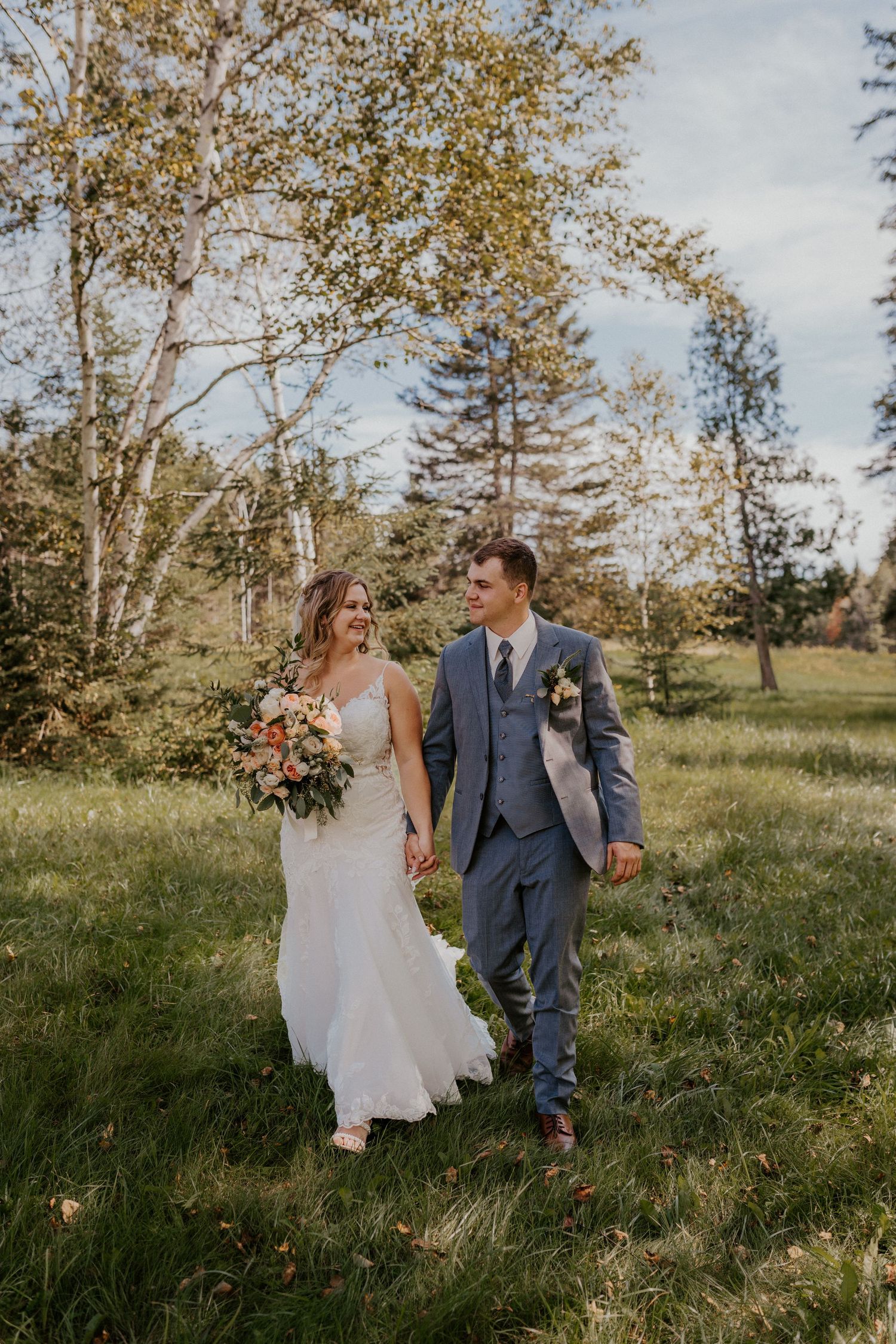 A couple runs playfully through a grassy field on an autumn day with golden trees in the background.