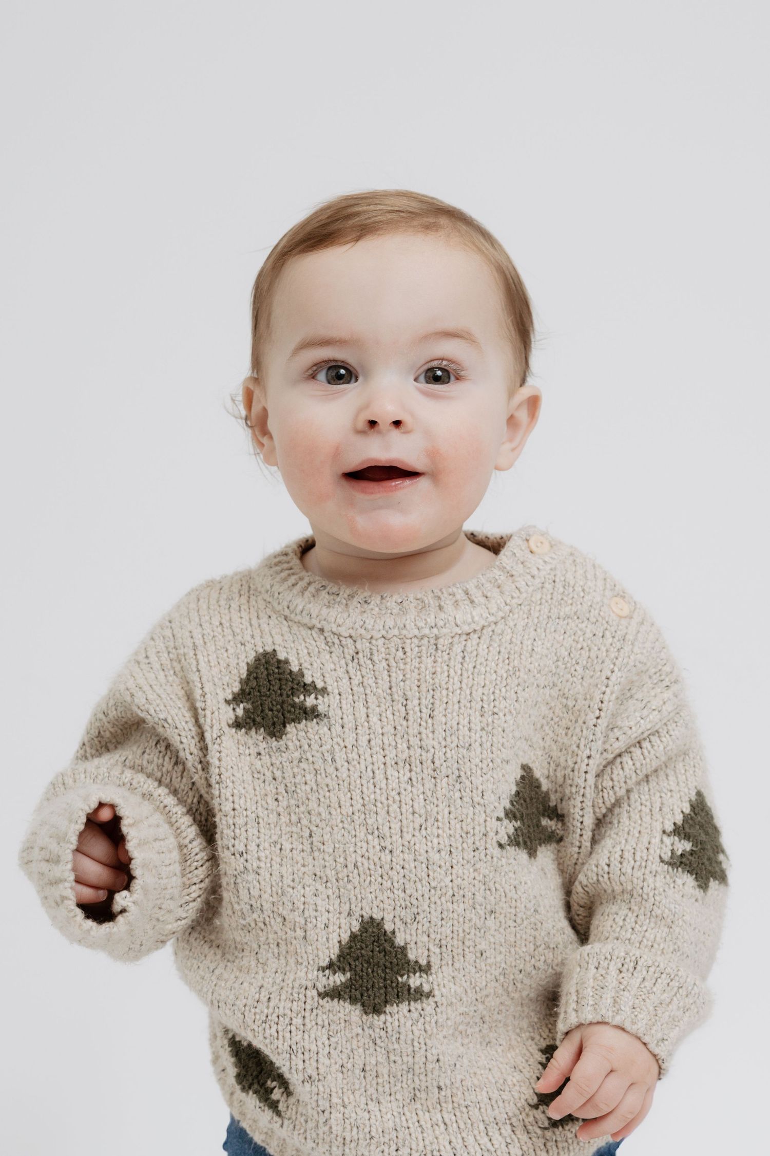 Baby wearing a cozy beige knit sweater with dark tree pattern design smiles at the camera.