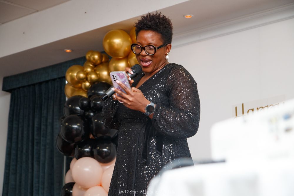 A person in a sparkly black dress holds a glass while celebrating at an elegant event with gold balloons.