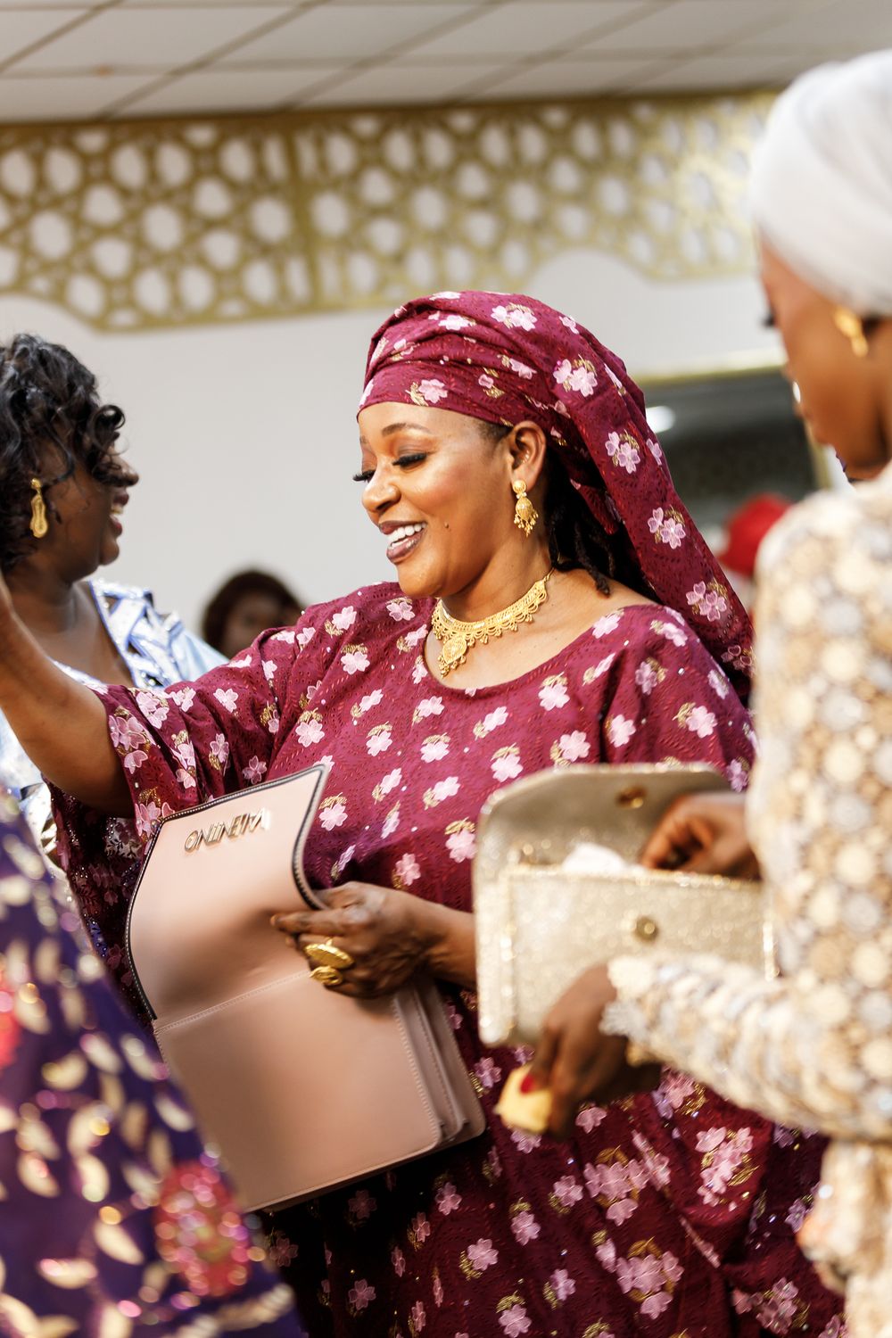 A woman in a burgundy floral headwrap and traditional attire smiles joyfully at a celebration event.