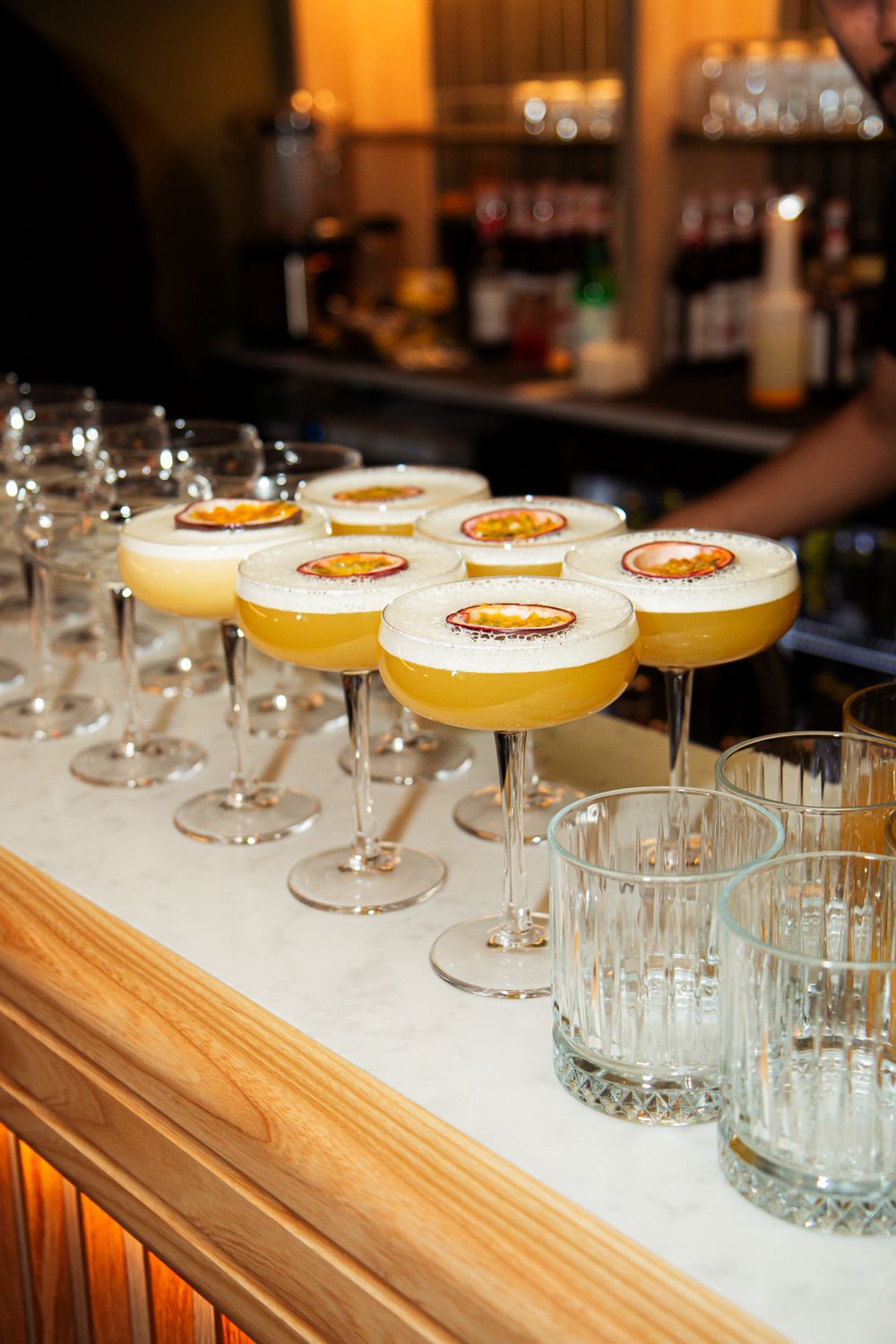 Several cocktail glasses filled with yellow drinks and garnishes are arranged in a row on a wooden bar counter.