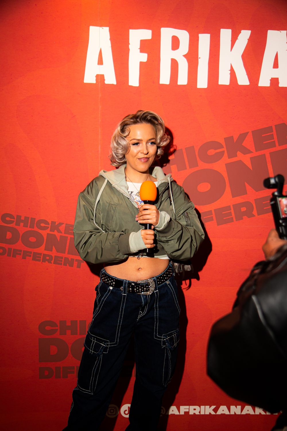 A performer in a crop top and baggy jeans holds a microphone against a red backdrop with 'AFRIKA' text.