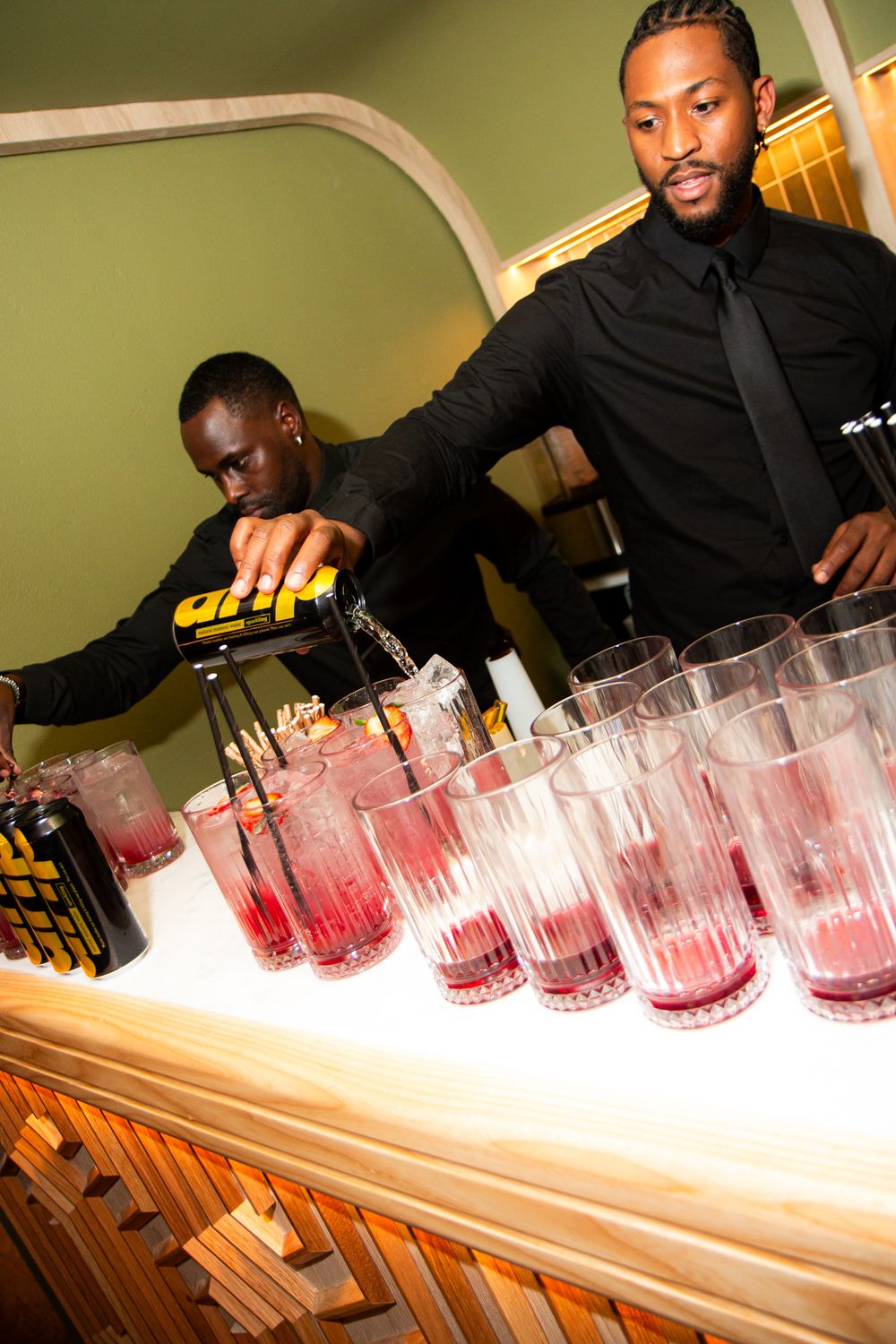 A bartender in black attire arranges a row of pink cocktails in glass tumblers on a wooden bar counter.