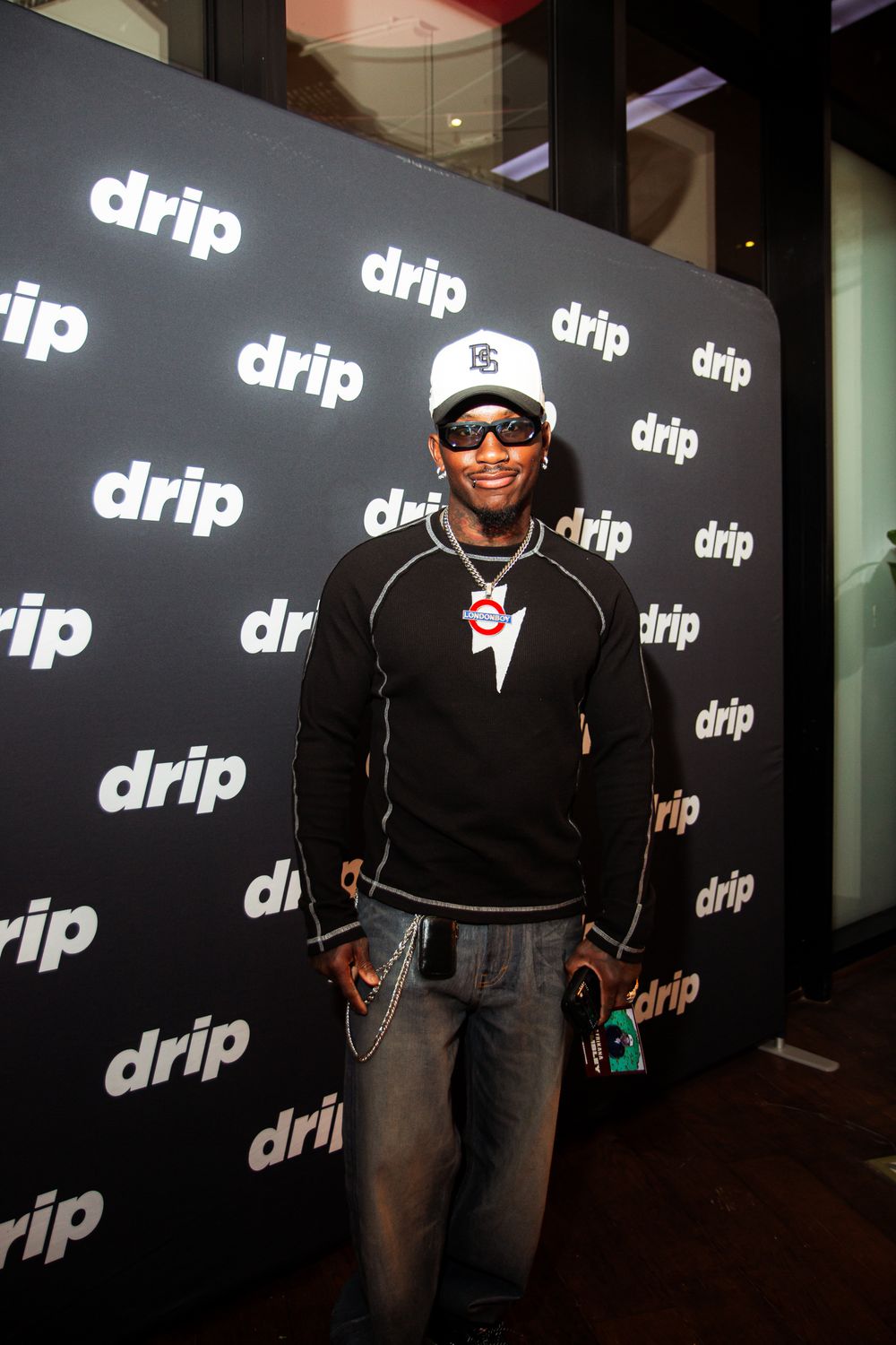 A person poses at a promotional event in front of a black backdrop with repeating white 'drip' logos.