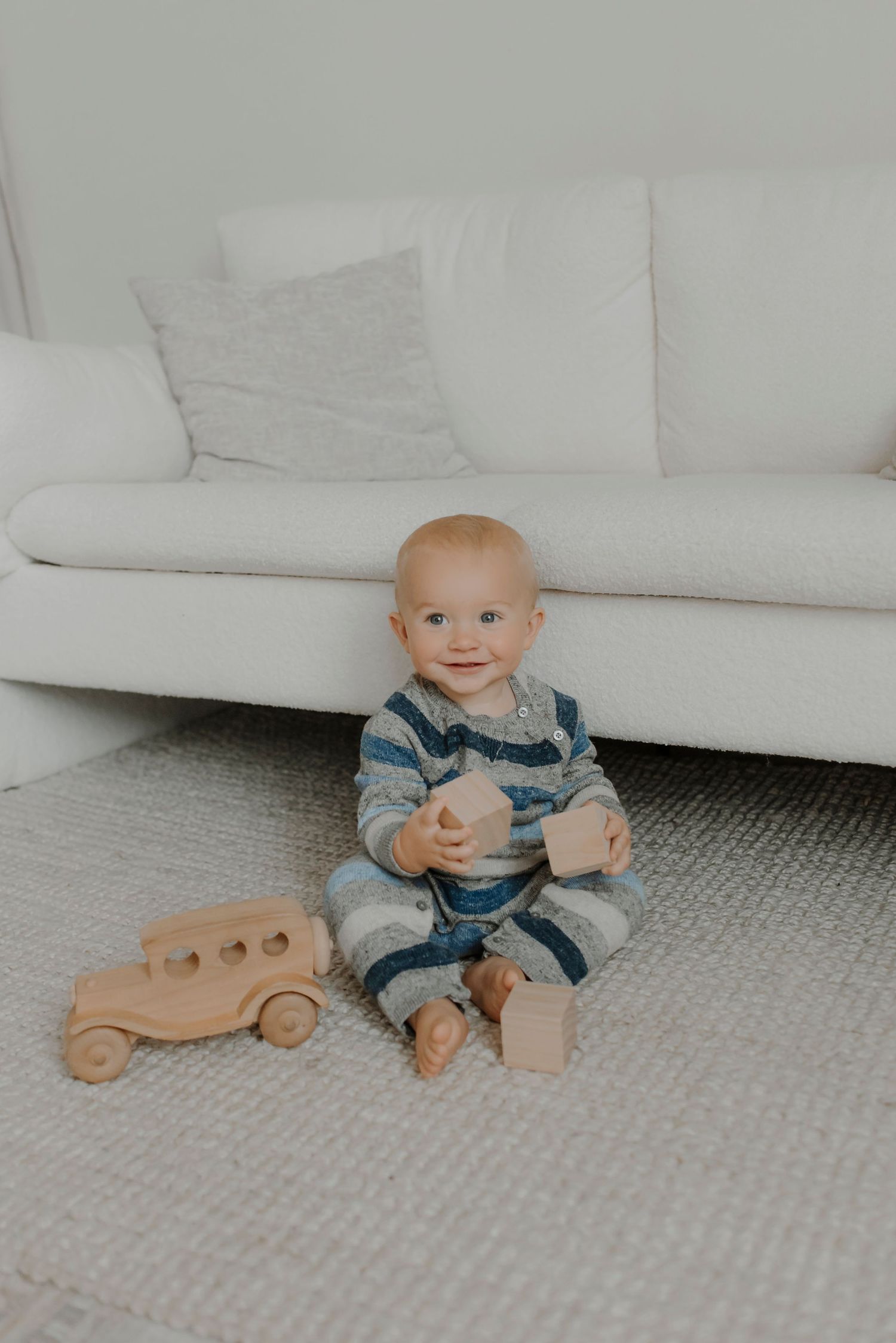 Baby playing with wooden toy car on carpet beside white couch.