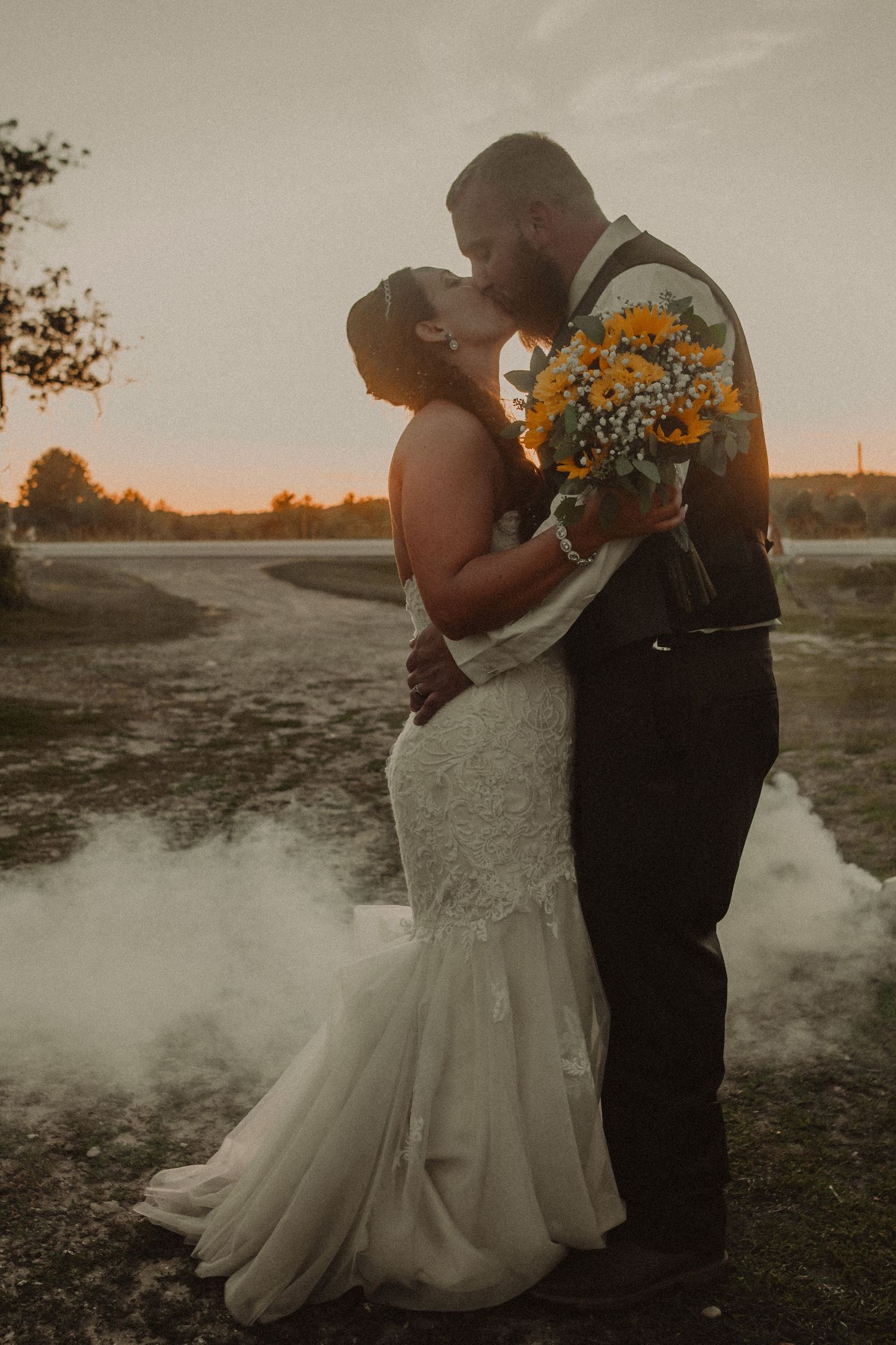 A romantic sunset wedding photo shows a couple embracing while holding a yellow bouquet, with misty ground and golden light.